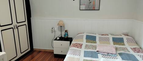 Une chambre avec un plancher en bois, un lit recouvert d’une courtepointe à motifs, une table de chevet avec une lampe, un tableau accroché au mur et une armoire.