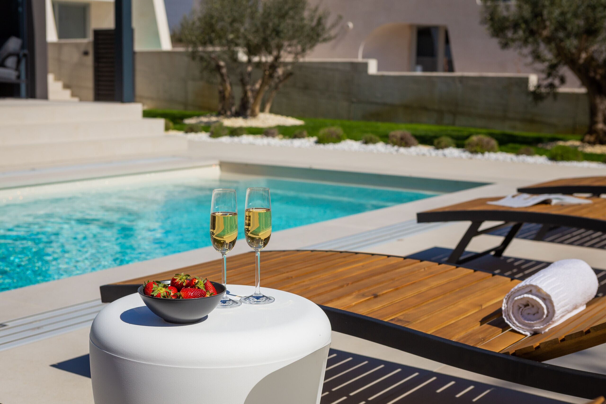 A poolside setting with a table holding a bowl of strawberries and two glasses of white wine, a wooden lounge chair, and a towel rolled up.
