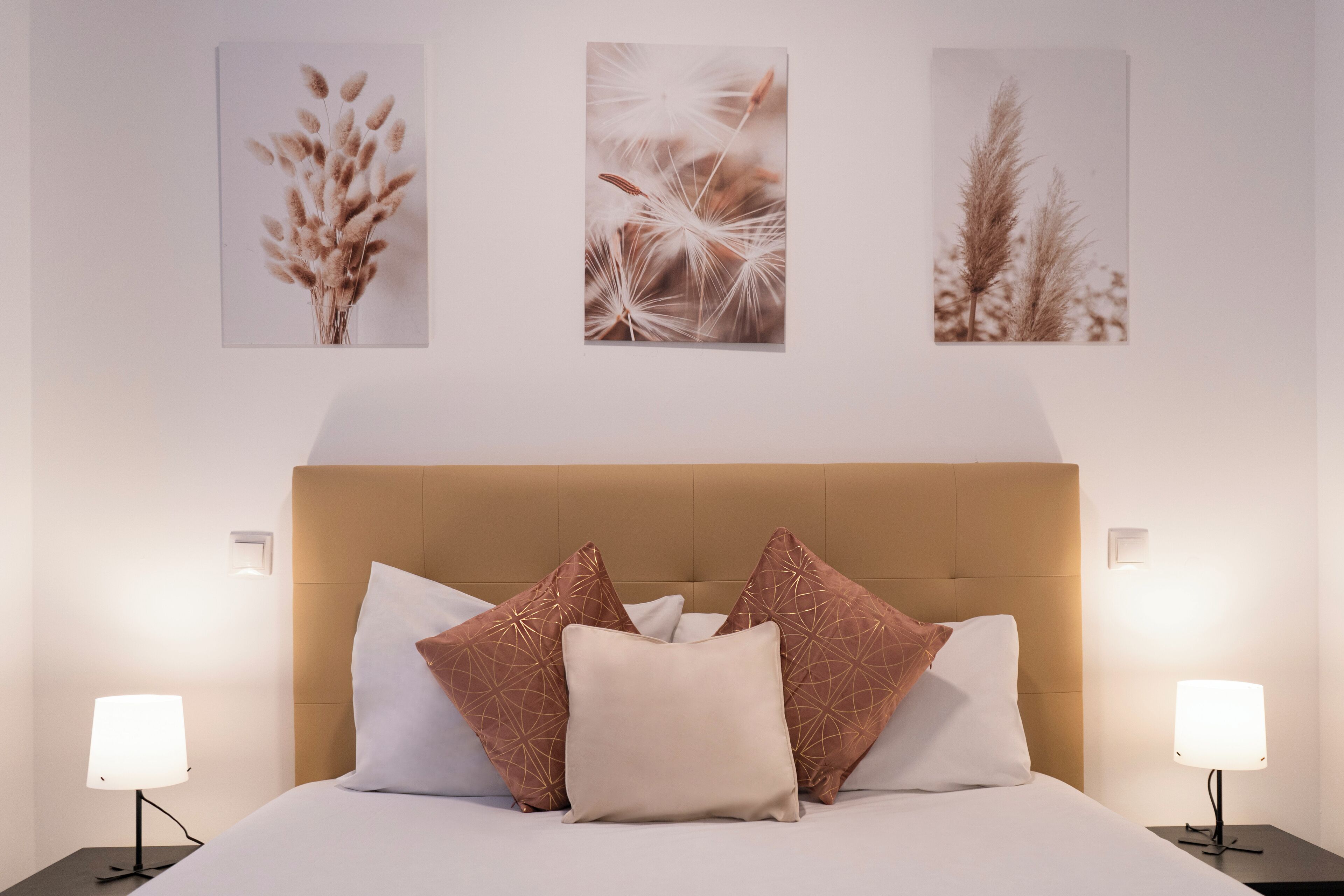 A neatly made bed with a beige headboard, two bedside lamps, and three framed botanical prints on the wall.