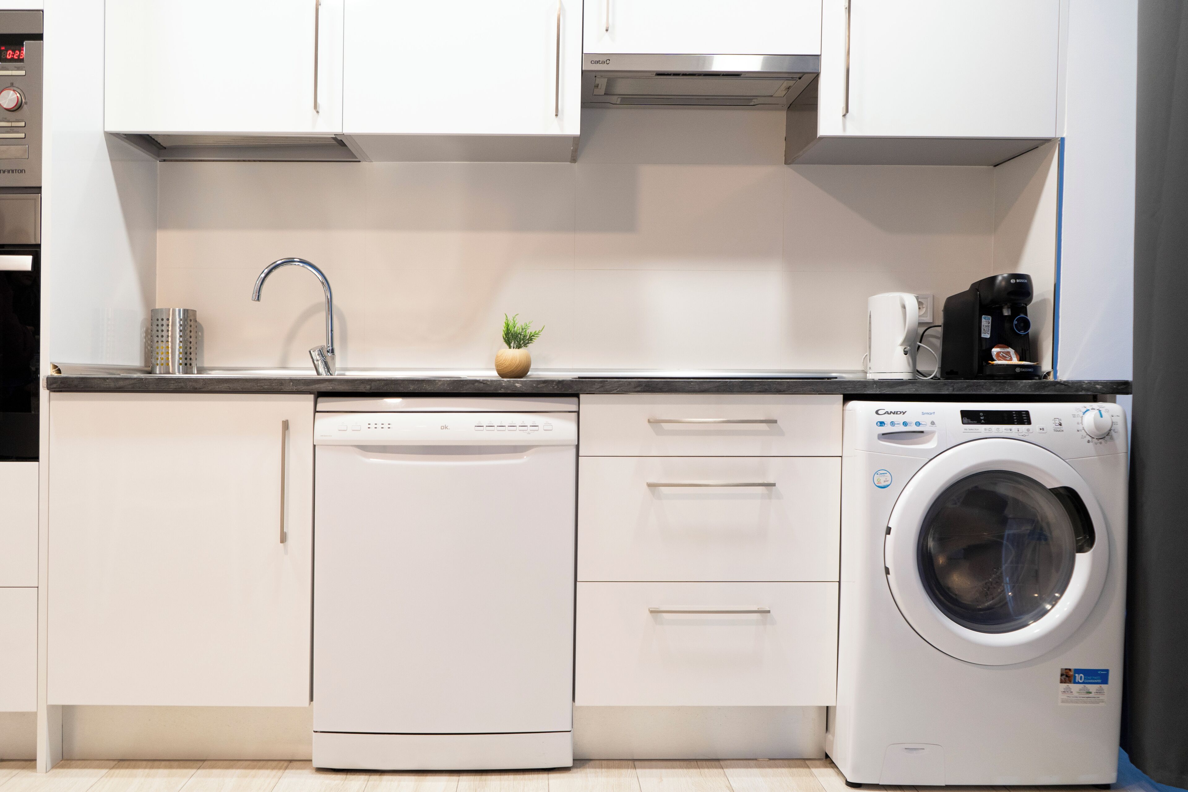 Cocina compacta con armarios blancos, lavavajillas, lavadora y cafetera.