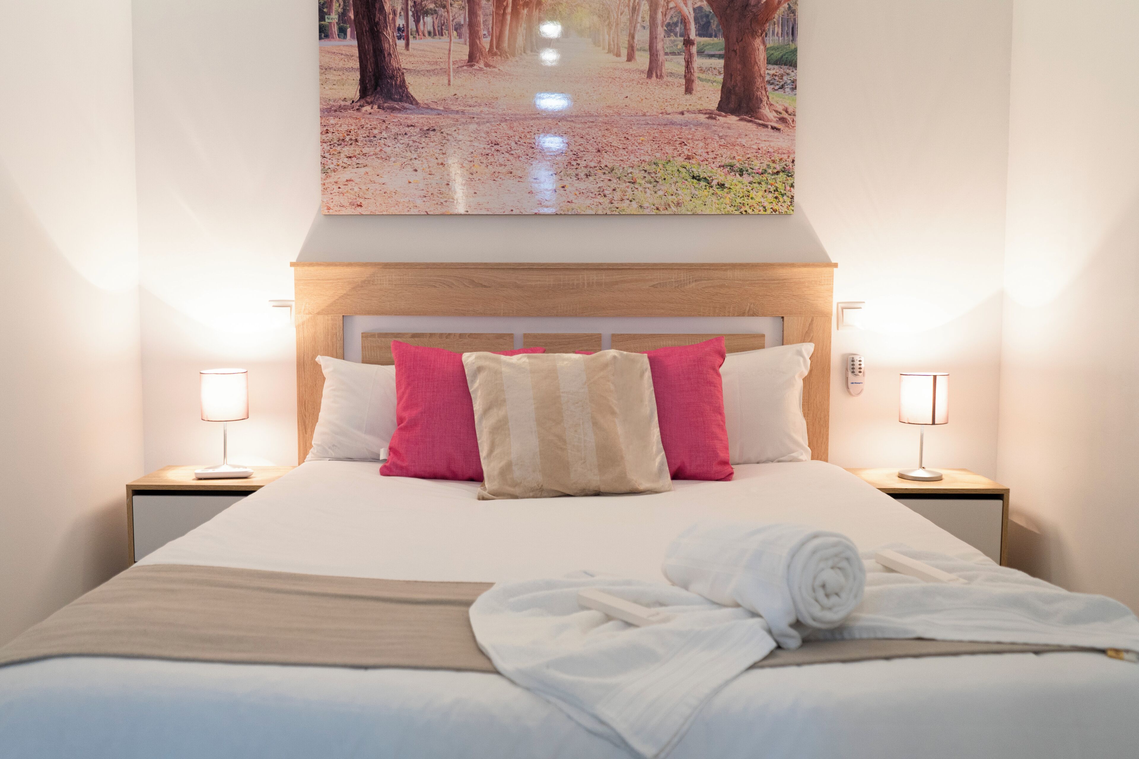 A neatly made bed with a white and beige striped pillow, a rolled white towel, and a wooden headboard. There are two bedside lamps on either side of the bed.
