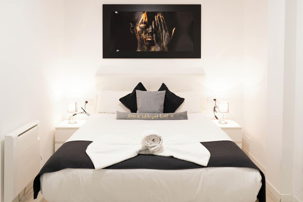 A neatly made bed with a white and black bedspread, a towel, and a pillow with a gray pillowcase. There is a framed artwork above the bed and a bedside table with a lamp on each side.
