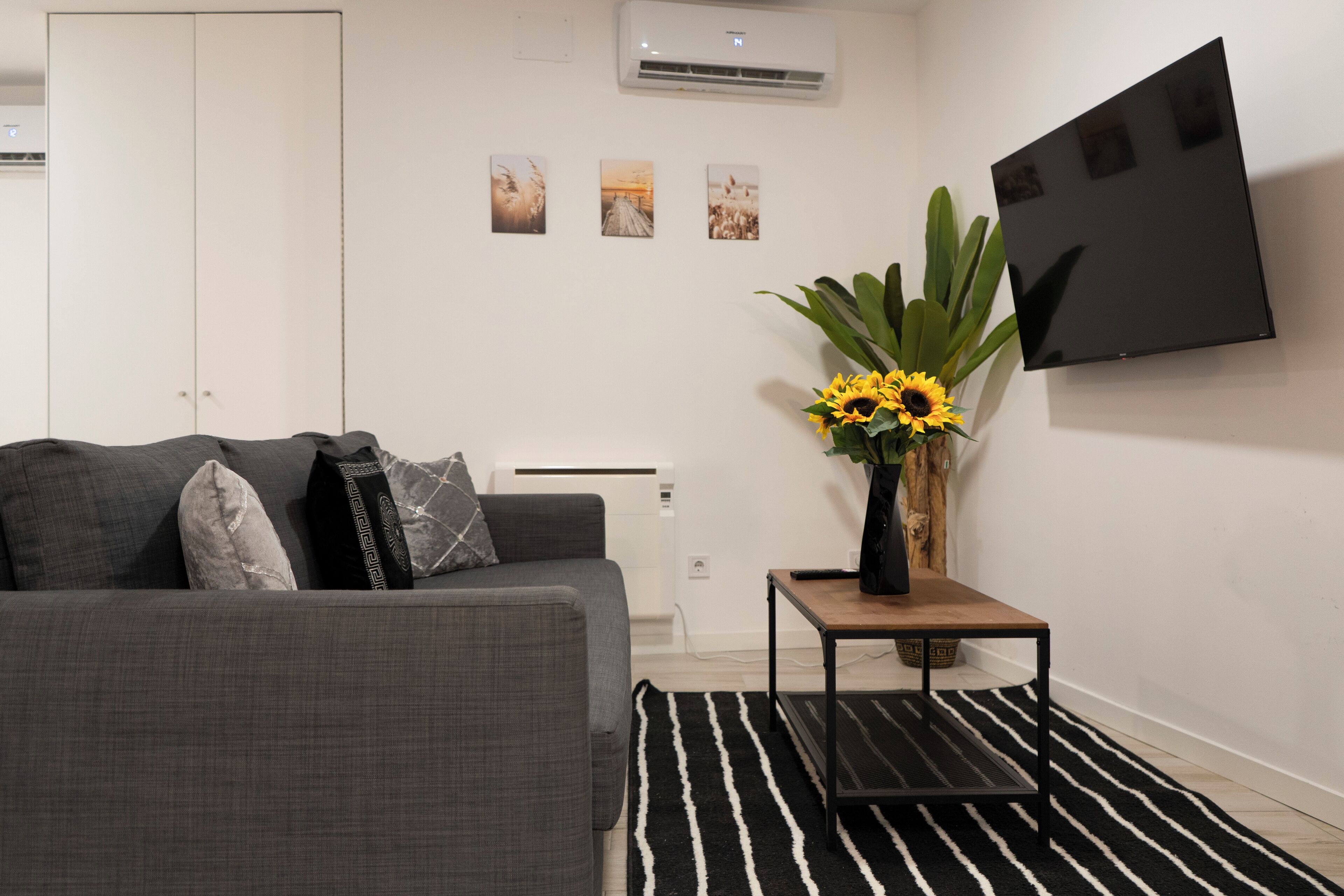 Un salon avec un canapé gris, un tapis rayé noir et blanc, une petite table d’appoint en bois sur laquelle est posé un vase rempli de tournesols, et un téléviseur à écran plat fixé au mur.