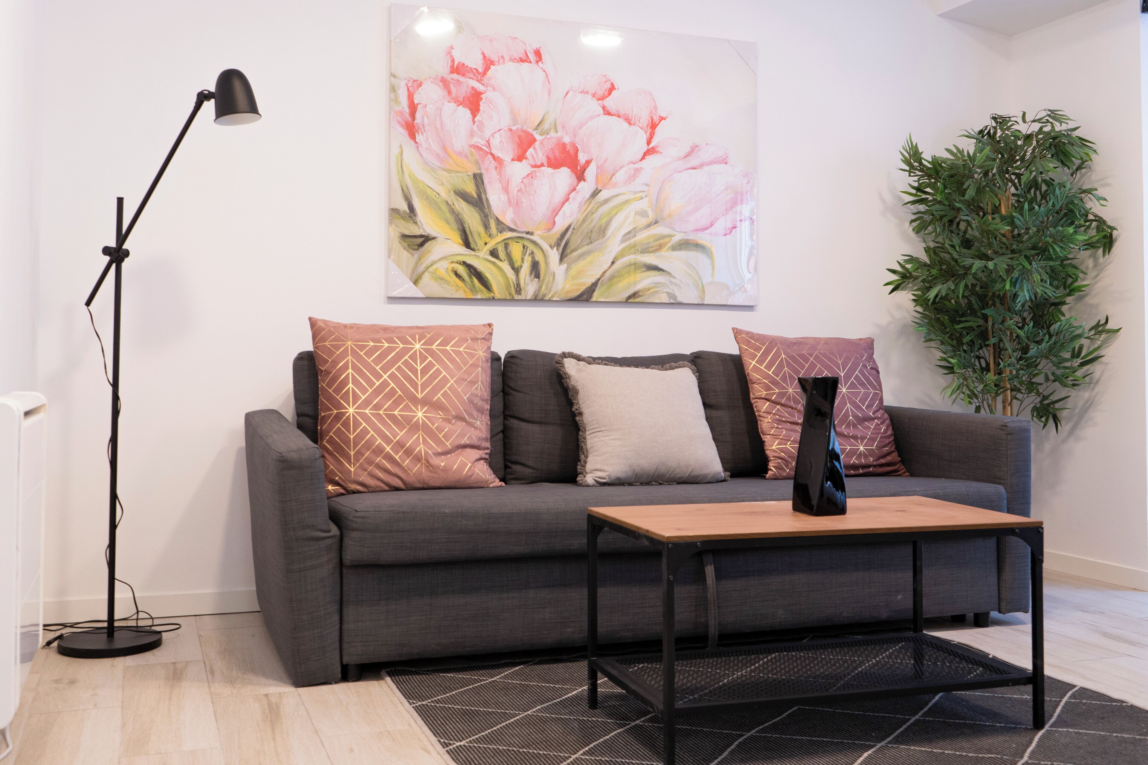 A modern living room with a grey sofa, patterned cushions, a wooden coffee table, a floor lamp, a large floral painting, and a green plant.