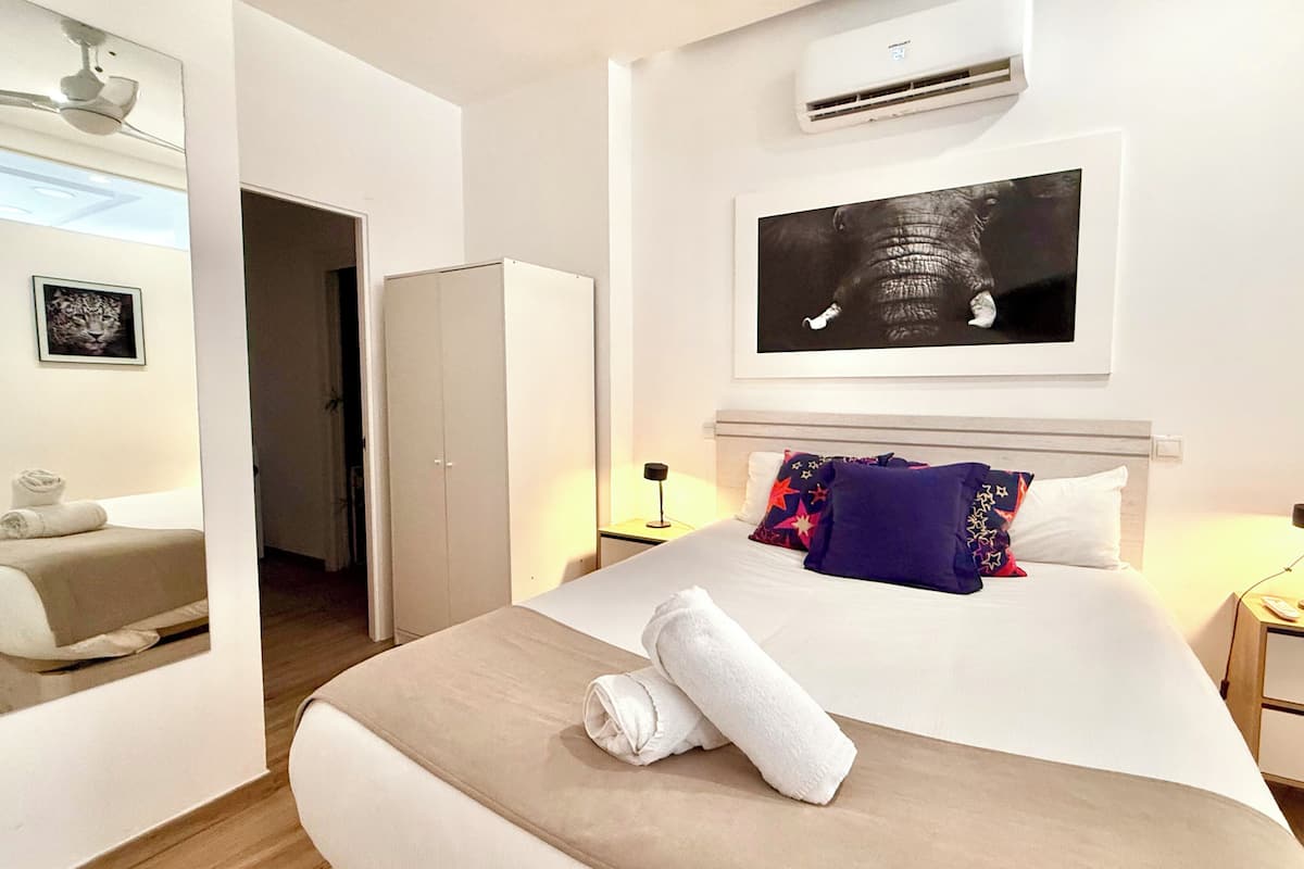 A hotel room with a bed, a nightstand, an air conditioning unit, a framed picture on the wall, and a door leading to another room.
