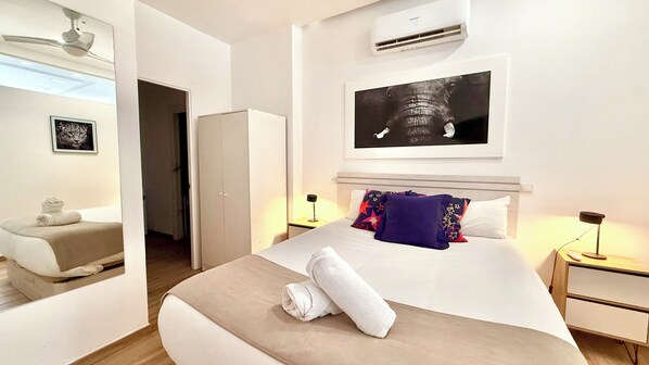 A hotel room with a bed, a nightstand, an air conditioning unit, a framed picture on the wall, and a door leading to another room.