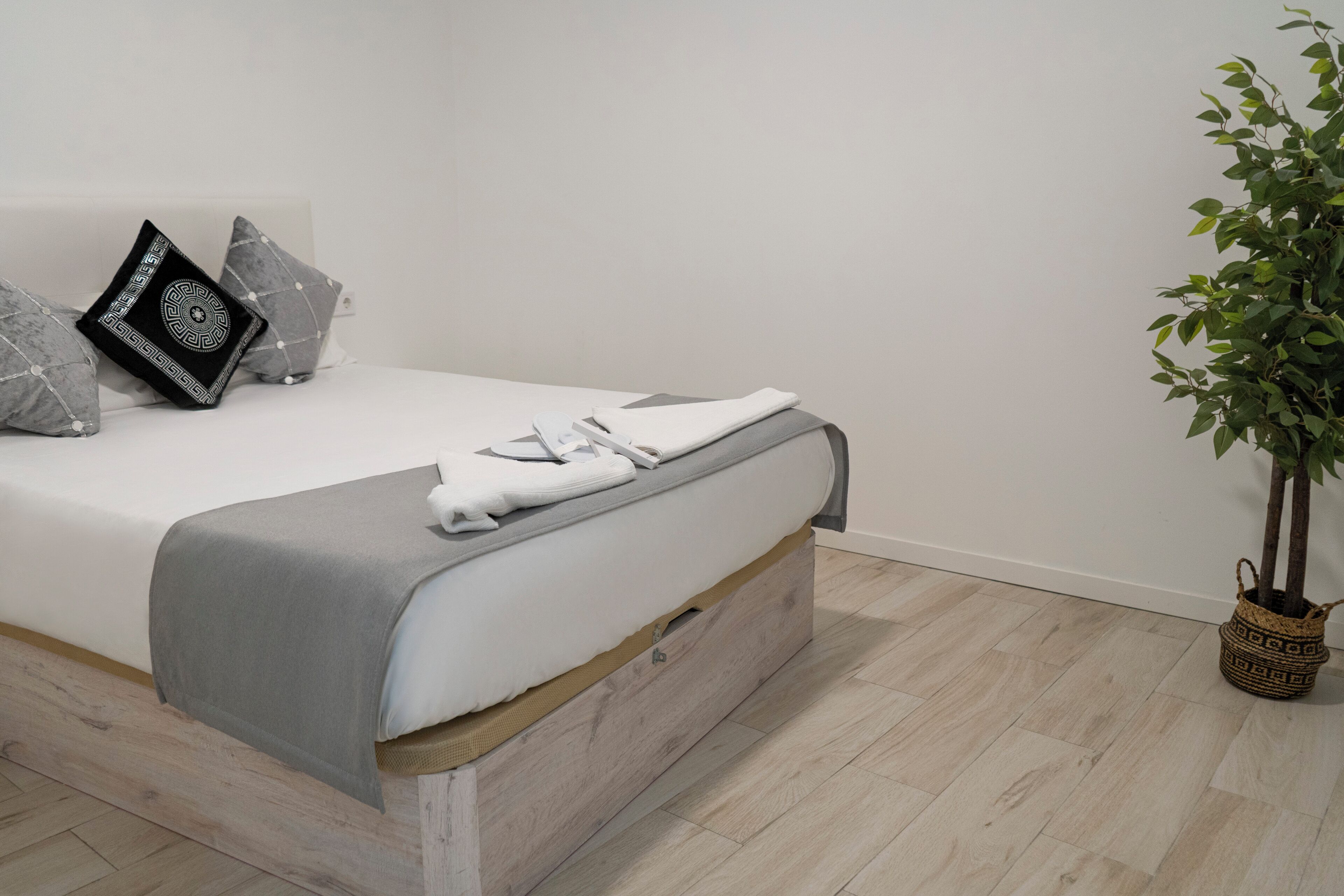 Una cama bien hecha con una manta y almohadas grises, una pequeña planta en una cesta tejida y un suelo de madera.