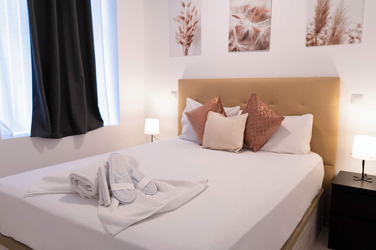 A neatly made bed with a towel and slippers, a bedside table with a lamp, and wall art above the bed.