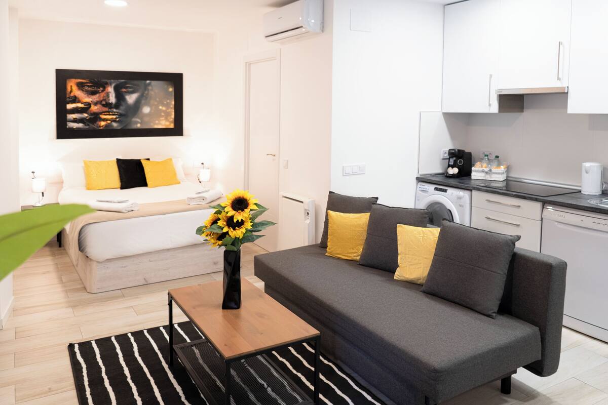 A modern apartment with a grey sofa, a bed with yellow pillows, a black and white striped rug, and a kitchen area with a washing machine.