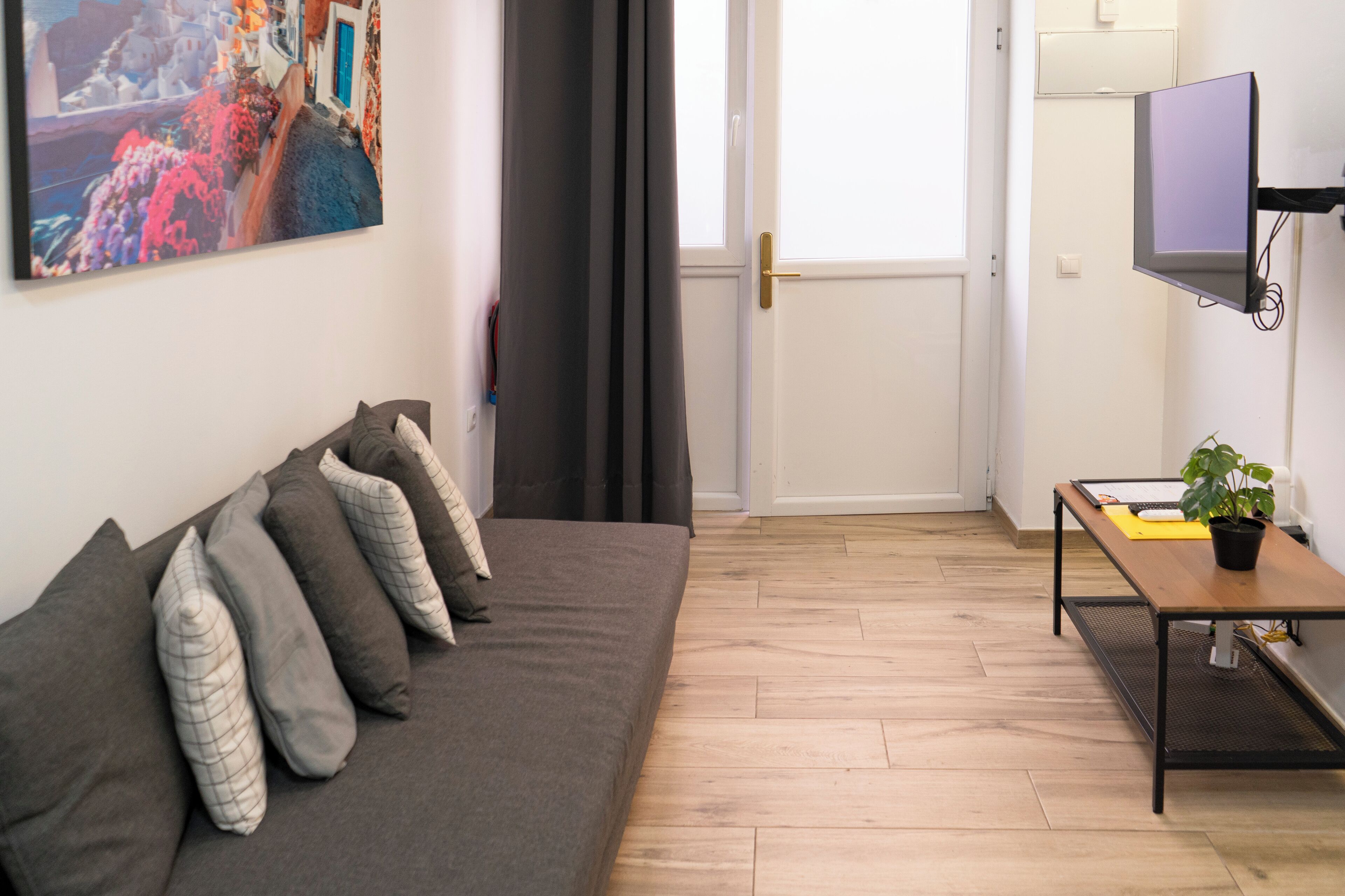 Una sala moderna con un sofá, un televisor de pantalla plana montado en la pared, una mesita con una planta y una puerta que da a otra habitación.