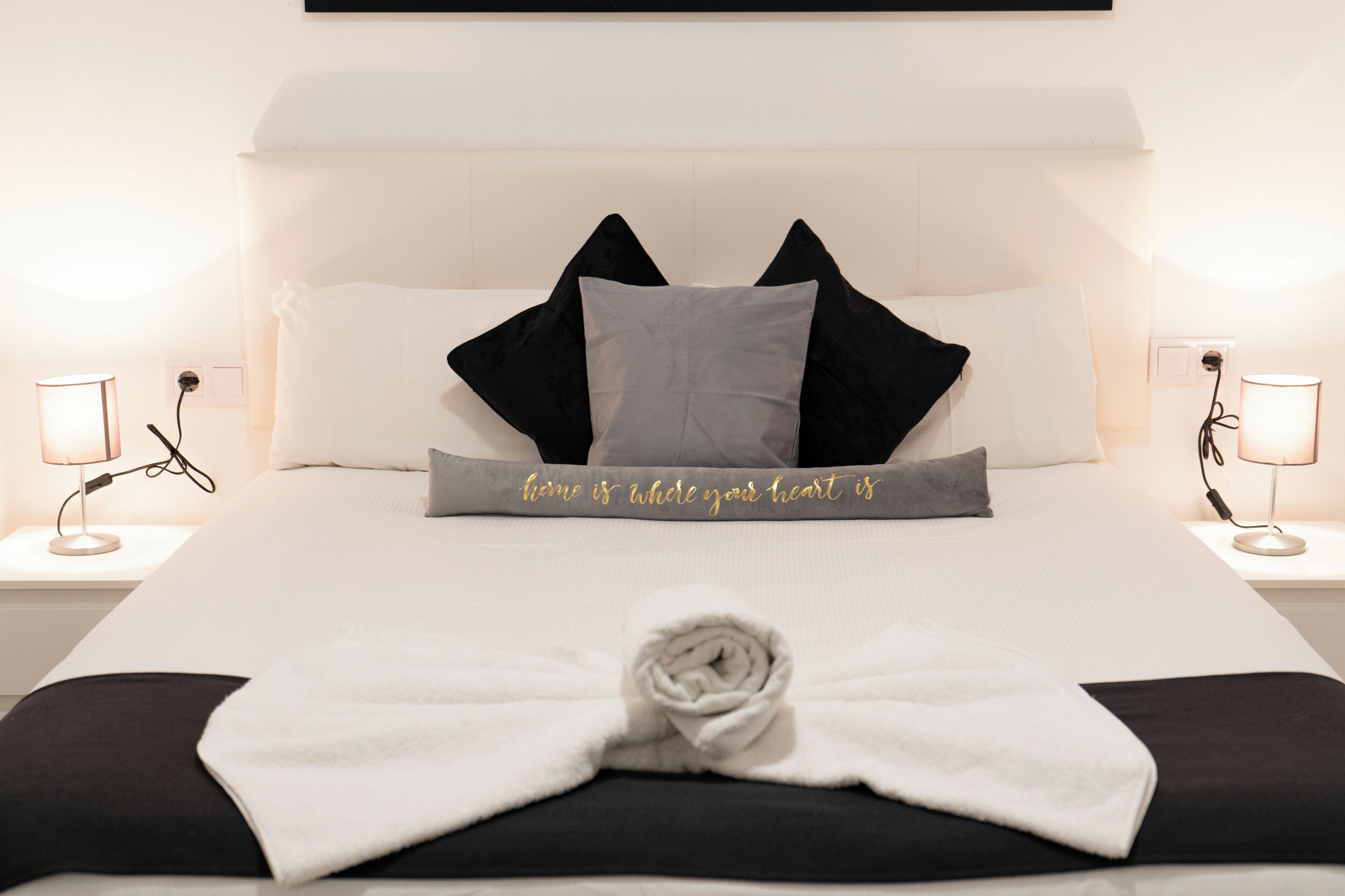 Una cama impecablemente arreglada con una toalla como adorno central, dos lámparas de noche y una almohada con una cita motivadora.