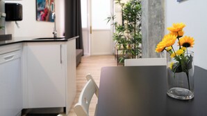 Moderni hotellihuone, jossa on ruokailutila. Tilaan kuuluu pöytä, jossa on maljakko täynnä keltaisia kukkia, valkoinen tuoli ja musta työtaso.