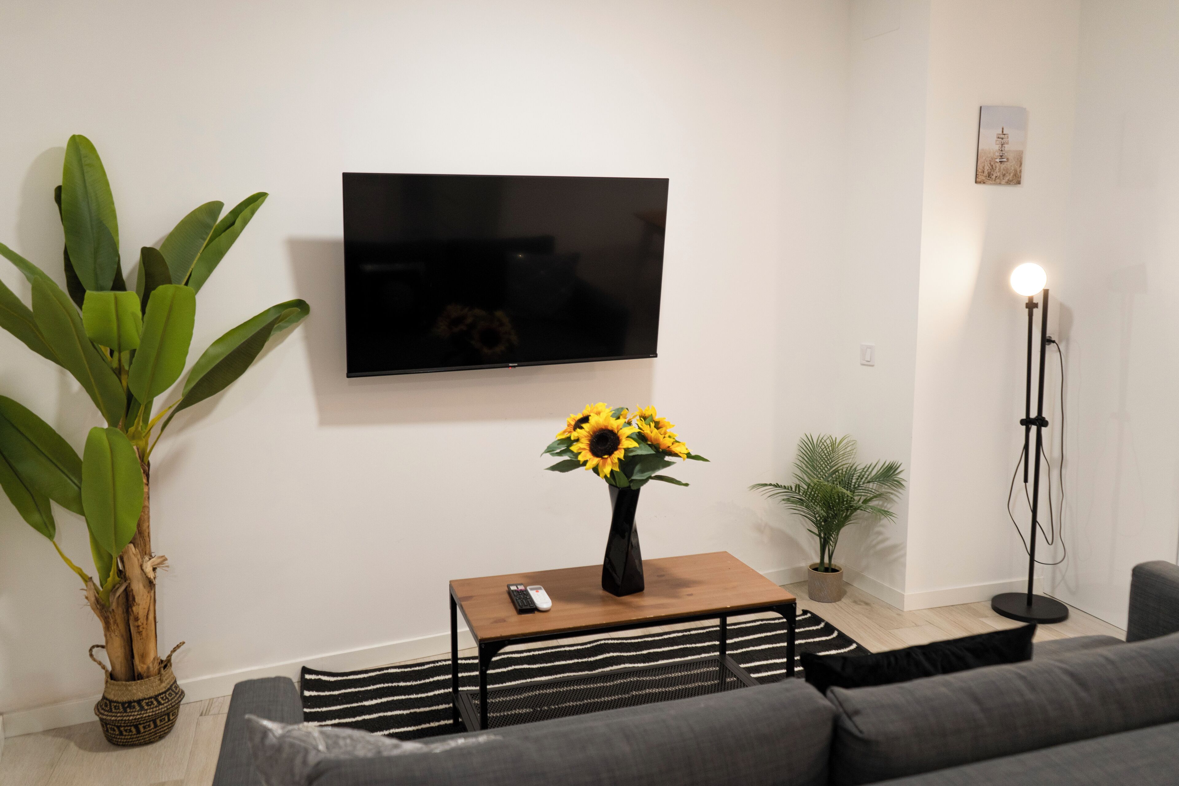 A living room with a flat-screen TV, a wooden coffee table with a vase of sunflowers, a grey sofa, a striped rug, and a tall floor lamp.