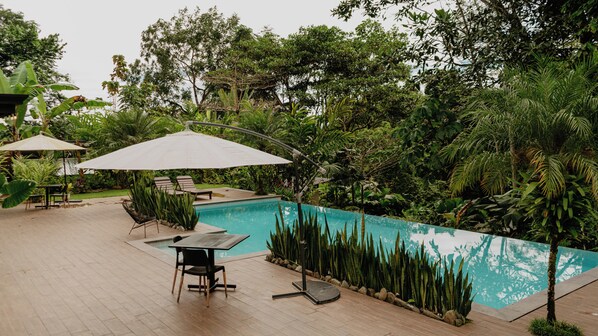 Zona junto a la piscina con mesa y sillas bajo sombrillas, rodeada de exuberante vegetación.