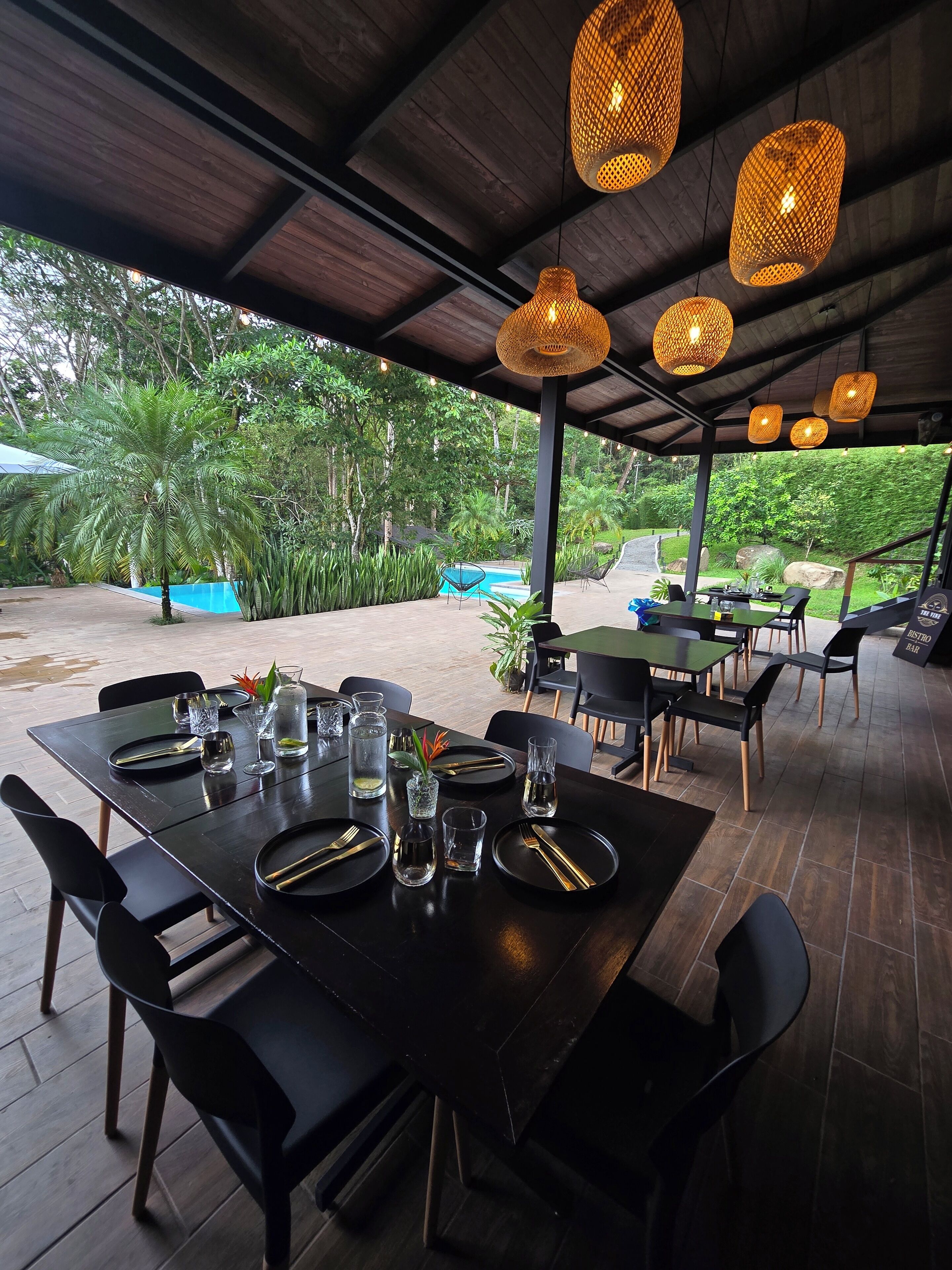 An outdoor dining area with tables set for two, surrounded by chairs, under a covered roof with hanging lights.