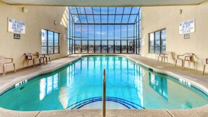 An indoor swimming pool with clear glass walls, chairs around the pool, and signs stating 'NO LIFEGUARD ON DUTY'.