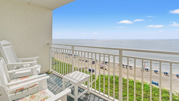 A balcony with white chairs and a small table, overlooking a beach with umbrellas and a clear sky.