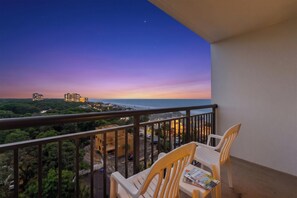 Un balcon donnant sur un coucher de soleil sur l’océan, une table avec des chaises et un magazine.