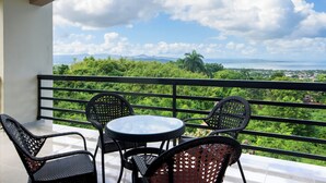 Ein Balkon mit rundem Tisch und vier Stühlen, der auf eine üppige grüne Landschaft und eine ferne Aussicht auf Berge und Meer blickt.