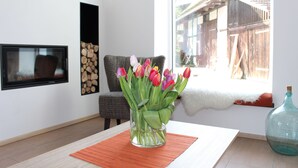 A modern living room with a glass vase of tulips on a wooden table, a fireplace, a chair, and a view of a wooden building outside. - House Apple Blossom - No 3 (Langenargen)