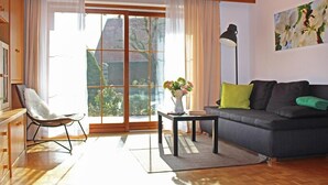 A living room with a sofa, a chair, a coffee table, and a floor lamp. There is a large window with curtains, and a painting on the wall. - House Apple Blossom - No 4 (Langenargen)