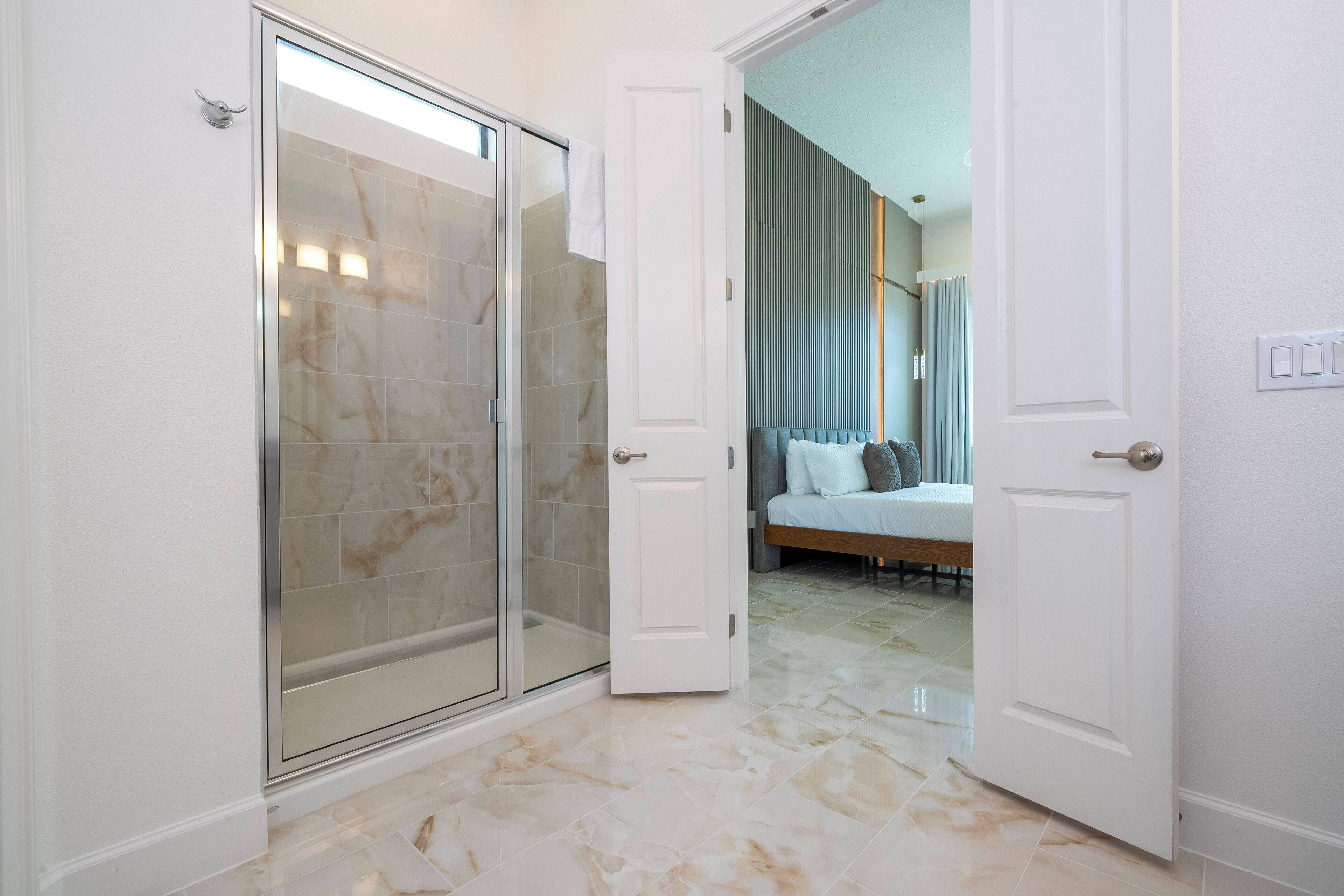 A bathroom with a glass shower enclosure, a white door leading to a bedroom with a bed and a nightstand.