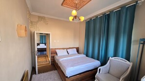 Un dormitorio con una cama de madera, una silla, un tapete estampado, una lámpara de techo y vistas a otra habitación a través de una puerta abierta.