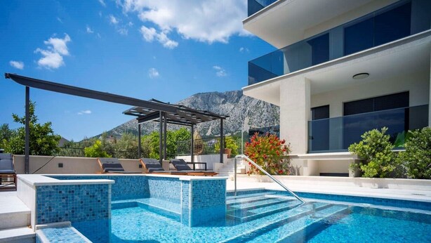 A modern outdoor swimming pool with a pergola, lounge chairs, and a mountain backdrop.