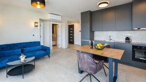 A modern apartment with a blue sofa, a small round table, a kitchen with dark cabinets, and a dining area with a wooden table and purple chair.