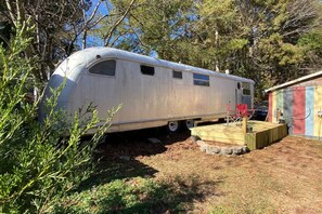 A vintage trailer with a small deck and a red chair, surrounded by trees.