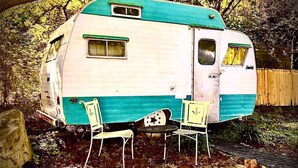 Uma caravana antiga com exterior azul-turquesa e branco, duas cadeiras de metal e uma pequena mesa na frente.