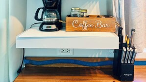 Una estación de café con cafetera, tazas y una caja de madera con la etiqueta 'Coffee Bar' sobre un estante blanco.