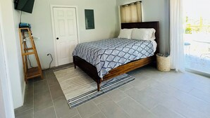 Un dormitorio con cama, ventilador de techo, televisor montado en la pared, puerta y ventana con cortinas.