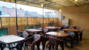 A cafe with multiple tables and chairs, large windows with a view of buildings, and a simple industrial-style ceiling. - Hotel Holy City (Amritsar)
