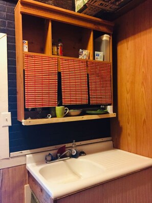 Eine kleine Küche mit einem Holzschrank über einem weißen Waschbecken, Regalen mit Geschirr und einem Fenster mit Jalousien.