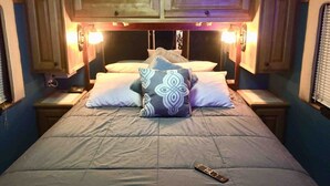 A bed with a remote control on it, wooden cabinets above, and two nightstands with lamps. - Stay Like a Rock Star! - Tour Bus in Durham, NC (Durham)