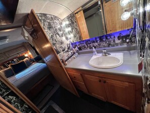 A compact RV interior with a kitchenette, a bed with a headboard, and a bathroom with a mirror and sink.