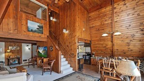Interior espacioso de una cabaña de madera, con escalera, comedor y sala de estar amueblada con sillas y mesas de madera.