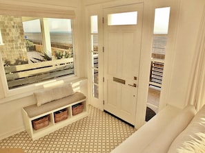 A room with a bench, wicker baskets, a patterned floor, and a door leading to a balcony with a view of the beach.