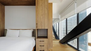Ein modernes Schlafzimmer mit einem großen Bett, einer Holztruhe und Blick auf eine Stadtlandschaft durch große Fenster.