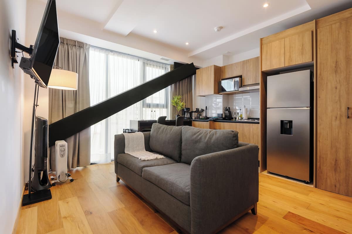 A modern living room with a sofa, a flat-screen TV, a standing fan, and a kitchen area with wooden cabinets and a refrigerator.