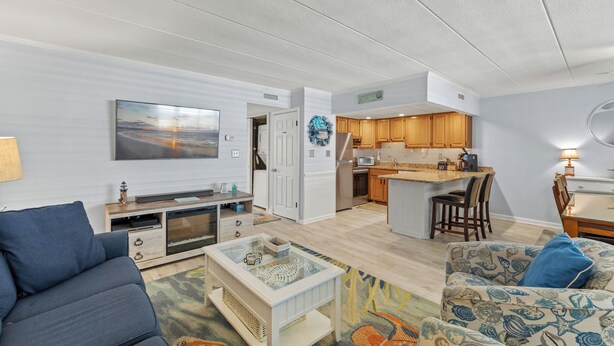 A modern living room with a sofa, a coffee table, and a TV. There is a kitchen area with wooden cabinets and a dining area with a table and chairs. - Canvas Back 202 by Seaside Vacations & Sales (Ocean City)