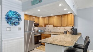 A modern kitchen with wooden cabinets, granite countertops, stainless steel appliances, and a decorative wall wreath. - Canvas Back 202 by Seaside Vacations & Sales (Ocean City)