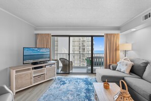 Un soggiorno moderno con televisione, divano, tavolino e vista sul mare da un balcone.