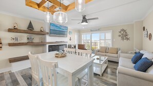 A spacious living room with a white sofa, a coffee table, and a dining area with a white table and chairs. There is a ceiling fan and pendant lights above the dining table.