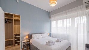 A bedroom with a bed, a nightstand, a lamp, a wardrobe, and a window with sheer curtains.