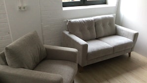 A room with a grey sofa and a matching armchair, a white brick wall, and a window with blinds.