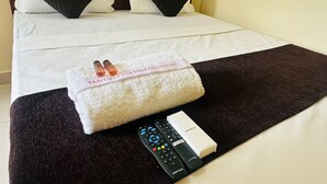 A neatly made bed with a white comforter, two black pillows, a white towel with a label, and a remote control on a dark brown bedspread. - S G R HOTEL & LODGE (Bengaluru)