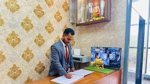 A man signing documents at a wooden reception desk with a computer monitor displaying surveillance footage. - S G R HOTEL & LODGE (Bengaluru)