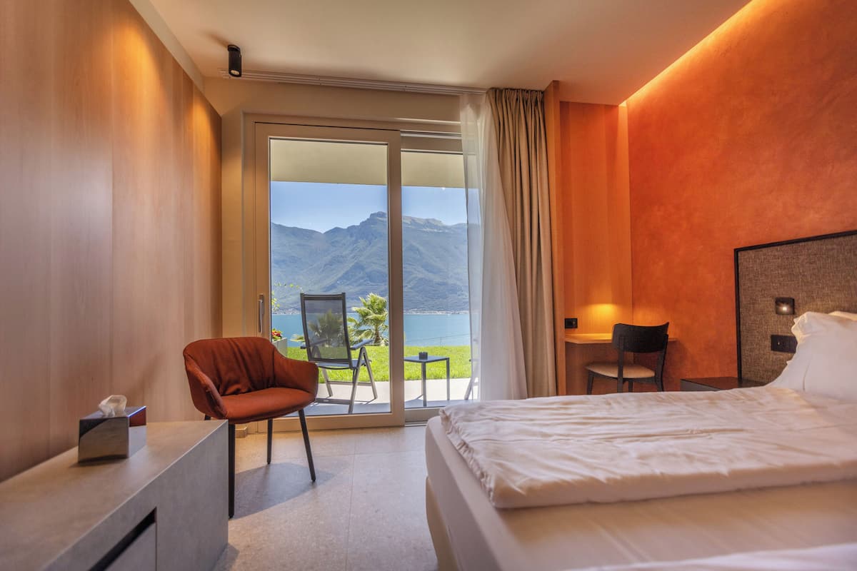 A hotel room with a bed, a desk, a chair, and a view of a mountain and lake through a glass door.